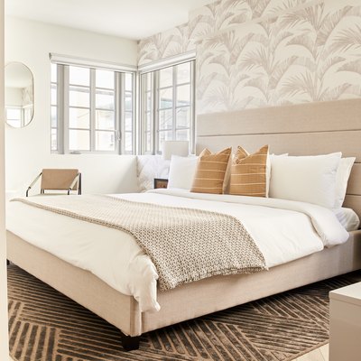 Modern bedroom with beige upholstered headboard, palm pattern wallpaper, nightstand, mirror, armchair, pillows, blankets, nightstand.